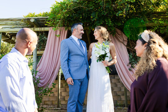 Vows starting, newlyweds exchanging under pergola draped pink, bride in white dress holding lilies
