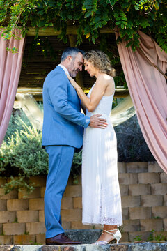 Couple standing on platform under pergola with pink drapes, vines, wearing blue suit and lace dress