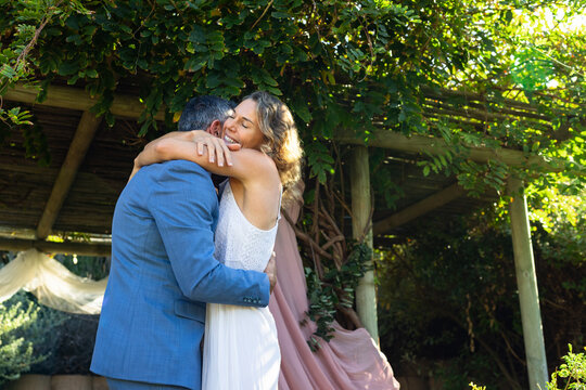 Couple embracing at garden pergola with pink drape, in white dress and blue suit, celebrating vows