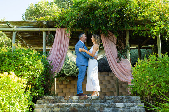 Couple embracing stone steps under garden pergola pink drapes, man in suit, woman in white dress