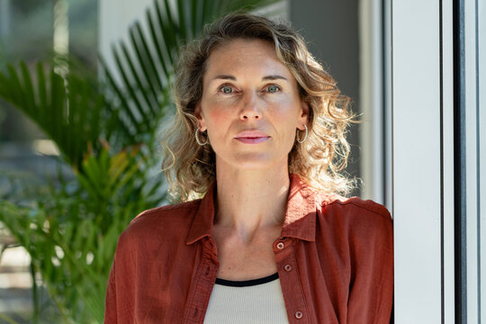Woman standing by white window frame, wearing rust shirt, ribbed tank, hoops, potted palm nearby