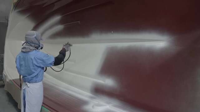 Medium shot of a worker in a respirator and hazmat suit spray painting the side of a massive fiberglass boat mold
