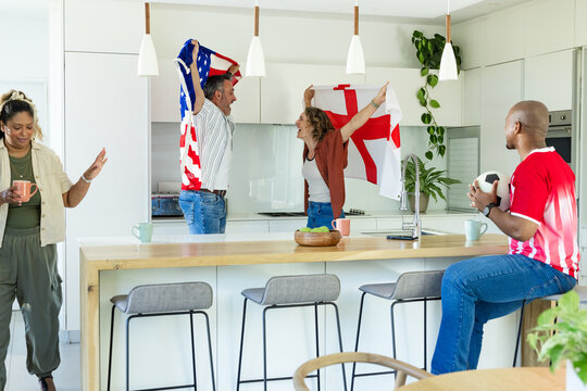 Diverse group cheering couple holding US ENG flags at kitchen island man red-jerseyed holding ball