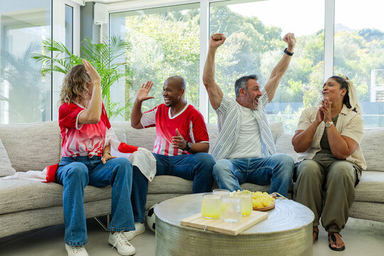 Diverse friends cheering, high-fiving after goal while sitting at home on sofa in red jerseys