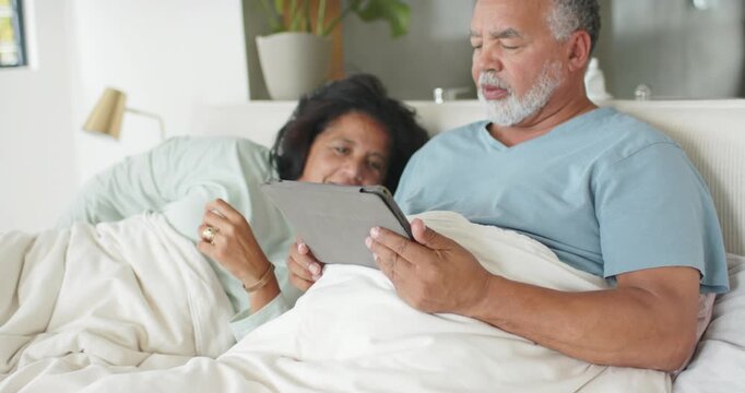 Senior African American couple watching tablet content, scrolling and leaning closer on white duvet