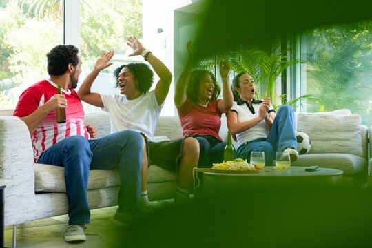 Diverse friends cheering and watching soccer match on sofa with chips, drinks, soccer ball