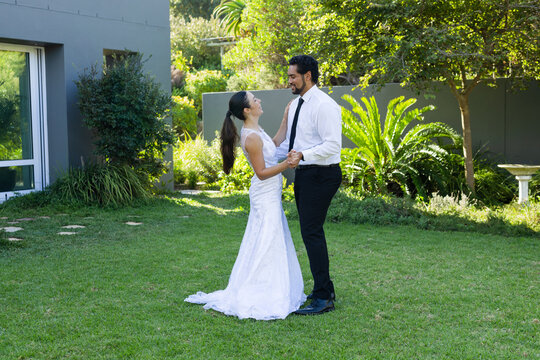 Diverse couple standing on lawn holding hands, smiling, wearing white lace dress and black necktie