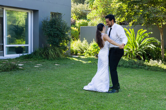 Couple dancing on well-kept garden lawn by modern house, holding hands, wedding gown and black tie