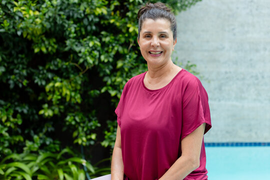 Mature woman wearing maroon top standing at pool edge with green wall and concrete wall, smiling