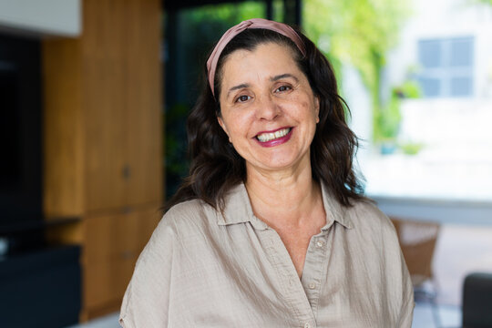 Woman standing smiling facing camera at home by wooden cabinets and glass door, wearing headband