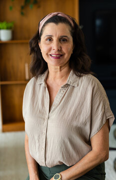 Mature woman standing in living room wearing beige blouse and pink headband, checking wristwatch