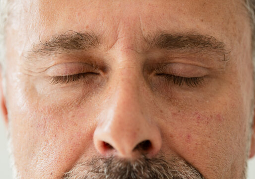 Mature male face filling frame, showing closed eyes, thick brows, large nose, stubble in studio