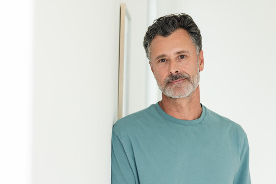 Mature male standing, leaning on framed panel in home studio, wearing teal crewneck shirt