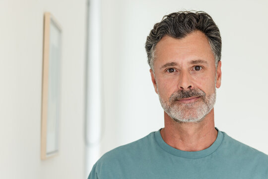 Middle-aged male standing by pale hallway wall wearing teal crew-neck T-shirt with framed art