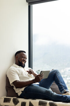 Adult man reclining on window seat in white shirt and jeans at home, holding tablet, laughing