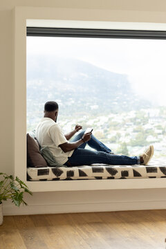 African adult man reclining at home in window seat wearing short-sleeve shirt holding phone, snack