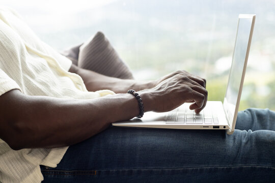 African American adult man typing on laptop on sofa by window wearing light shirt jeans bracelet