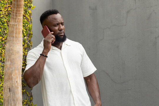 Adult African man standing by wall with vines, in white shirt, beaded bracelet, holding red phone