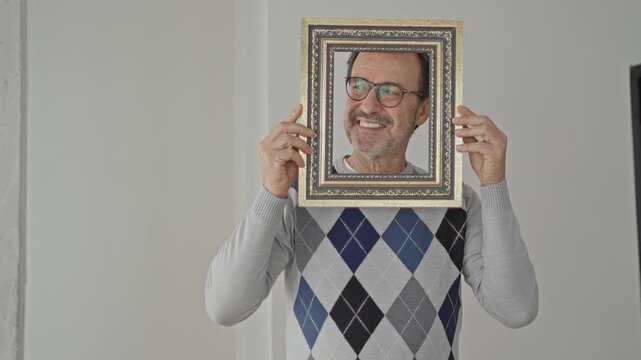 Man holds ornate picture frame over face, smiling and wearing an argyle sweater in a building interior hall; self reflection joy.