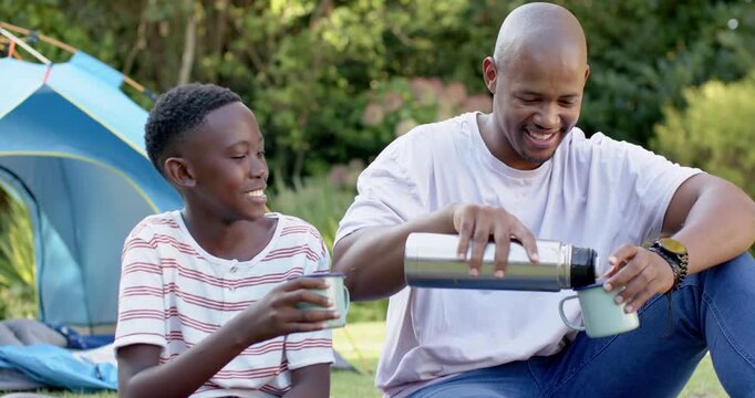 African American father and youth son by tent father opening thermos pouring mugs clinking bonding