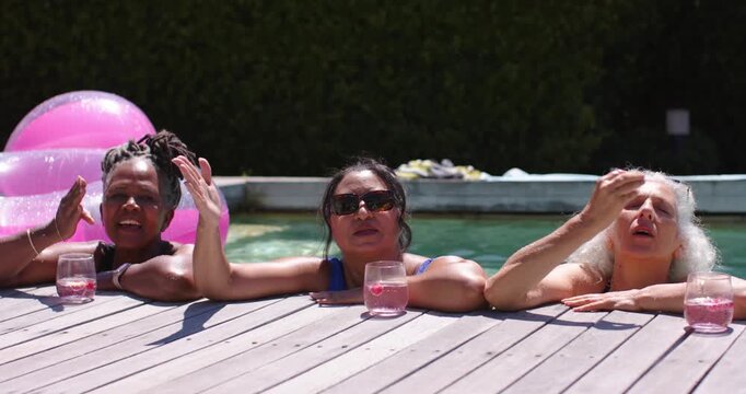 Diverse female friends talking and leaning on wooden deck at backyard pool, sipping pink drinks