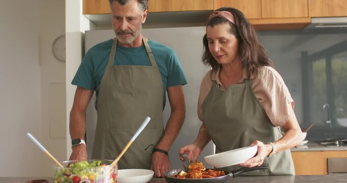 Mature couple in olive-green aprons plating at kitchen counter after cooking in skillet, serving
