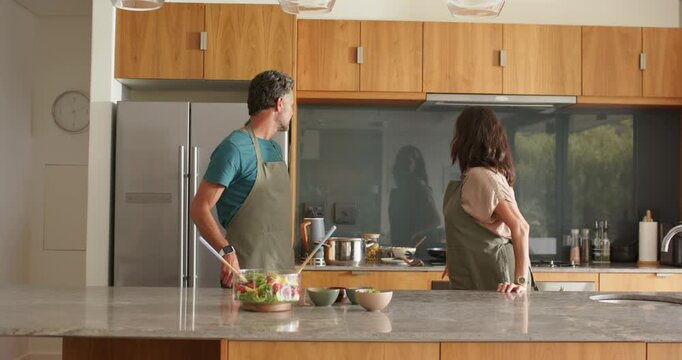 Mature couple wearing aprons touching arm then noticing reflection in glass and moving toward stove
