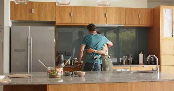 Senior couple in aprons making salad at island, man reaching behind her and embracing, sharing meal
