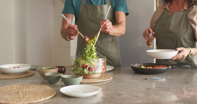 Couple in olive aprons at kitchen counter preparing dinner tossing salad with servers ladling pasta