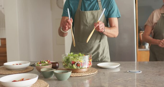 Diverse couple in aprons at kitchen island man picking salad servers tossing salad woman serving