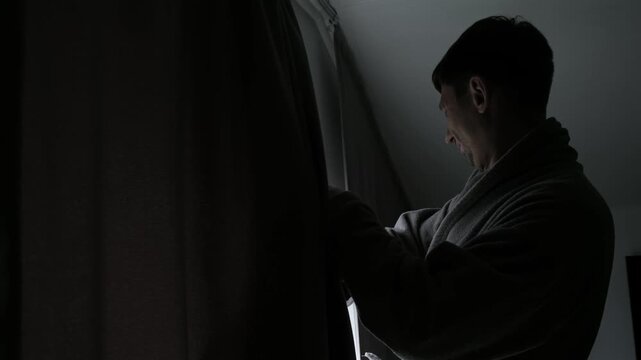 A man pulls back the curtains on the window and blocks out the light on a gloomy morning a concept of hangover, depression, photophobia, burnout, and a lack of joy in life.