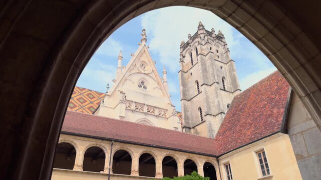 View of a historic gothic church courtyard in France framed by a stone arch. Elegant architectural composition with tower, arches, and tiled roofs. Atmospheric European landmark, ideal for travel and 