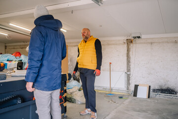 Fototapeta premium Two men discussing renovation plans inside a garage workspace, one wearing a yellow vest and the other in a blue jacket, with tools and materials visible in the background