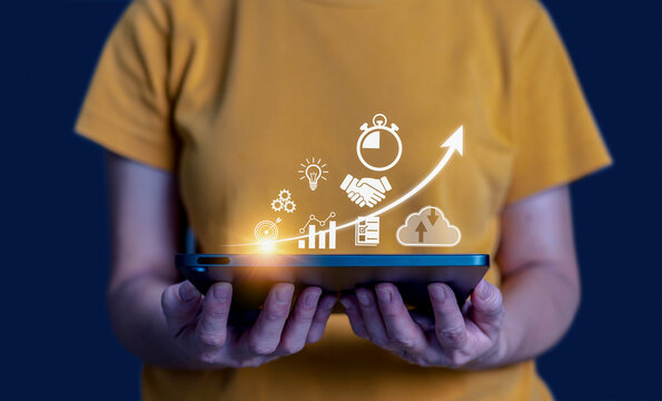 A woman holding a tablet with glowing virtual business icons&mdash;goal, handshake, gear, graph, and cloud&mdash;symbolizing growth, strategy, collaboration, and digital transformation.