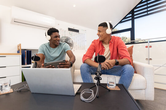 Recording Diverse male friends sitting on couch in home studio, wearing headphones and using laptop