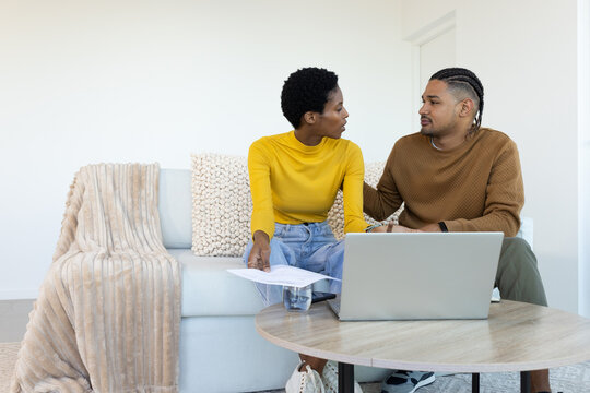Diverse couple discussing documents on living room sofa, with laptop and glass of water