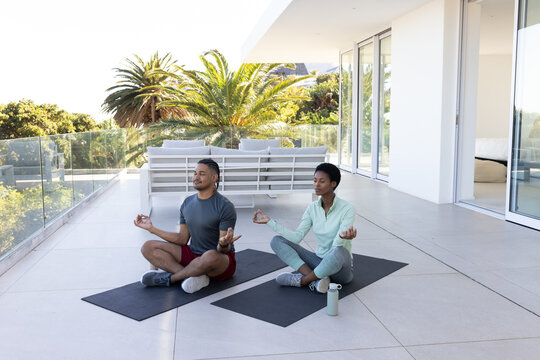 Meditating diverse couple sitting on black yoga mats on modern home terrace, with water bottle