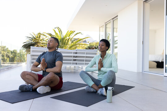 Meditating diverse couple on home patio by glass railing, with yoga mats, water bottle