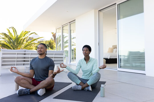 Meditating Diverse couple sitting cross-legged on terrace, with black yoga mats and water bottle