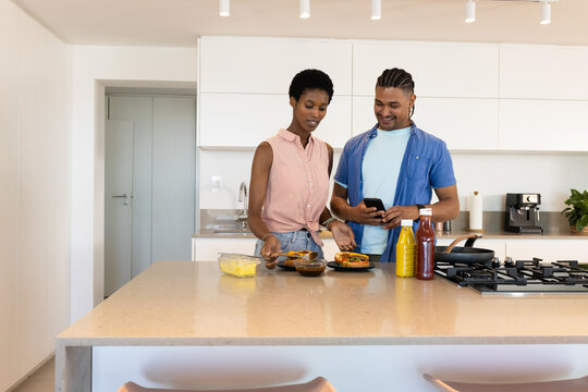 Diverse couple plating scrambled eggs in modern kitchen, with spatula and smartphone