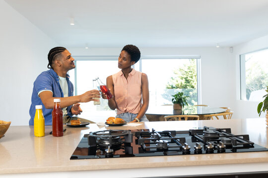 Toasting couple standing at marble island in home kitchen, with burgers, condiments and drinks