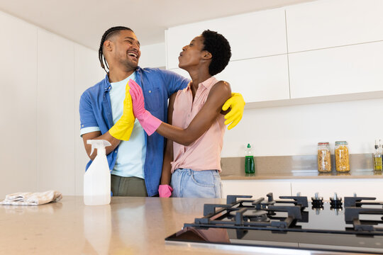 Laughing Diverse couple wearing rubber gloves cleaning countertop in kitchen, with spray bottle