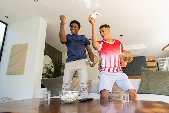 Celebrating diverse male friends raising controllers in living room, with popcorn bowl and water