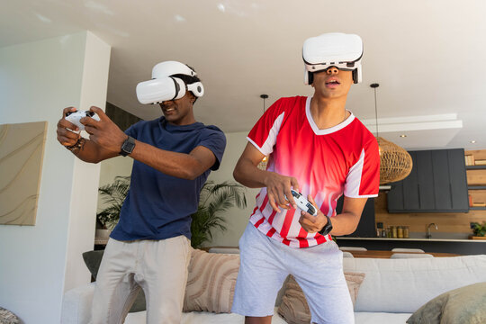 Playing Diverse male friends leaning into VR game in living room, with VR controllers and headsets