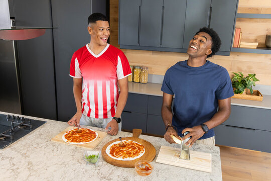 Preparing pizza on marble counter in kitchen by diverse male friends, using pizza peels and grater