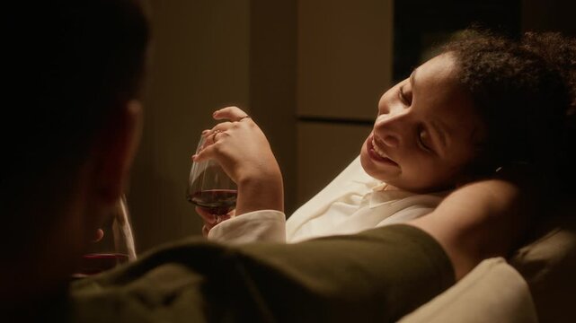 Over shoulder view of curly haired young woman leaning on husbands hand while chatting with him and drinking red wine