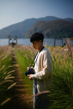 A young Asian man stands in tall grass by a lake, holding a camera and enjoying a peaceful outdoor moment surrounded by mountains