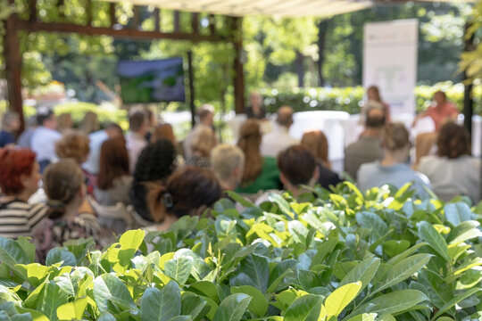 Outdoor business conference with professional audience attending presentation in green garden venue under canopy, sustainable corporate event setting