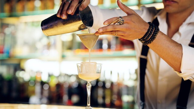 Bartender carefully pouring a freshly mixed cocktail from a shaker through a strainer into a glass with a large ice cube
