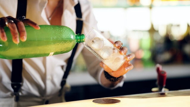 Bartender's hands preparing a beverage, pouring liquid from a green bottle into a glass filled with round ice, creating a refreshing drink